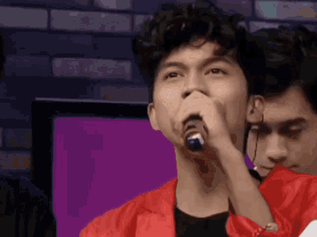 a man in a red jacket is singing into a microphone while another man looks on