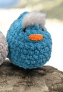 a close up of a blue crocheted bird with a white head and orange beak sitting on a rock .