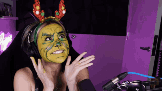 a woman wearing headphones and a headband with reindeer antlers has a green and yellow face painted on her face