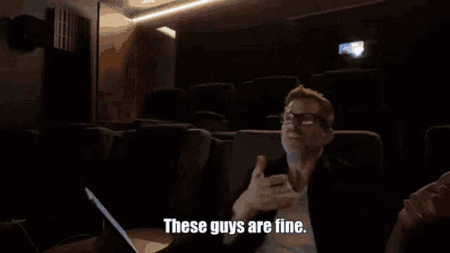 a man sitting in a dark auditorium says " these guys are fine " in front of a laptop