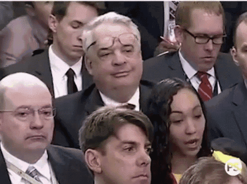 a group of people in suits and ties are sitting in a room and one of them is wearing glasses .