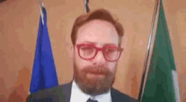 a man with a beard wearing glasses and a suit is standing in front of two flags .