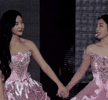 two women in pink dresses hold hands in front of a black background