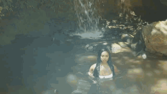 a woman in a white tank top is swimming in a stream near a waterfall