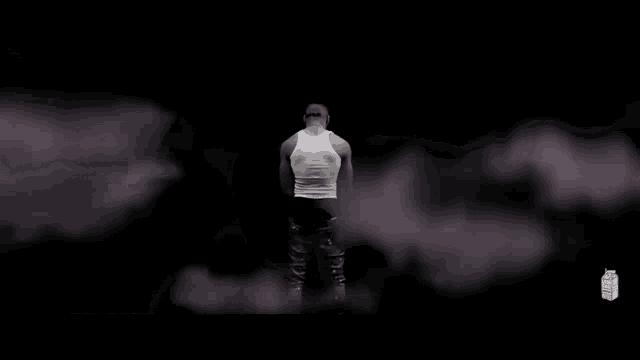 a man in a white tank top is standing in a dark room surrounded by purple smoke .