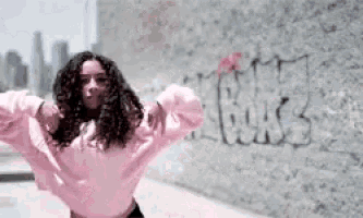 a woman in a pink sweater is dancing in front of a wall with graffiti on it