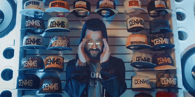 a man wearing glasses stands in front of a wall full of dennis hats