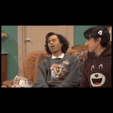 two men are sitting on a couch and one has a sweatshirt with a cat on the front