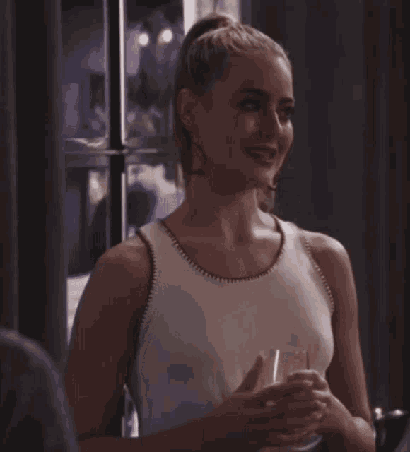 a woman in a white tank top smiles while holding a glass