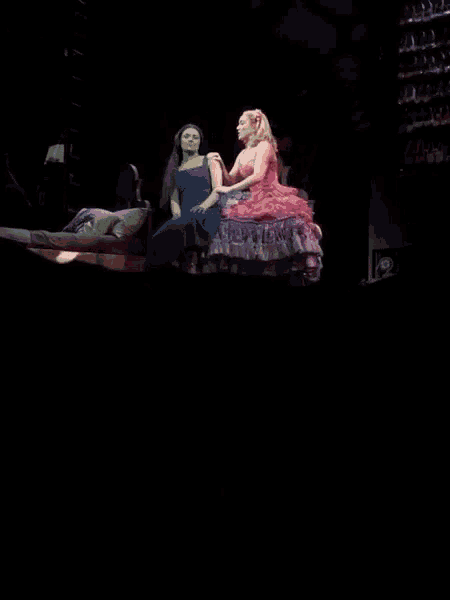 a woman in a pink dress is standing next to a woman sitting on a bed