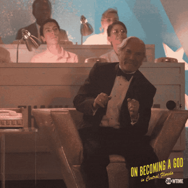 a man in a tuxedo sits in front of a sign that says " on becoming a god "