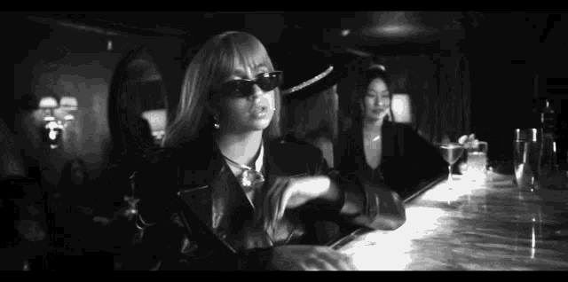 a black and white photo of a woman wearing sunglasses sitting at a bar