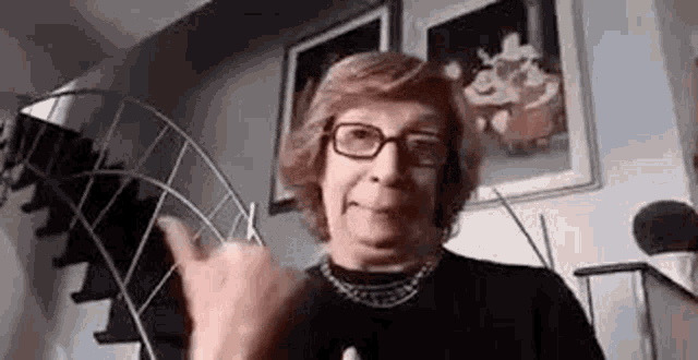 an elderly woman wearing glasses and a necklace is making a devil horns sign .