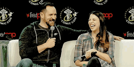 a man and a woman are sitting on a couch and laughing in front of an emerald city comic con banner