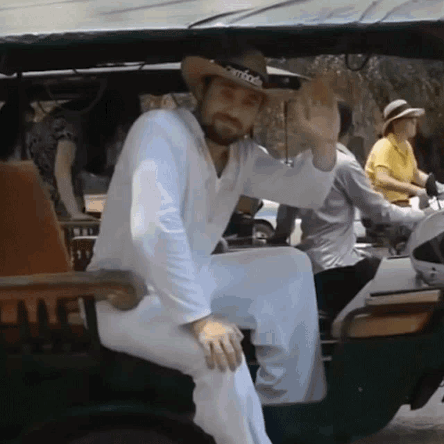 a man wearing a cowboy hat and white pants waves while sitting in a vehicle