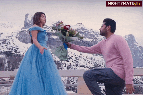 a man in a pink sweater is kneeling down to give a woman a bouquet of flowers in front of snowy mountains