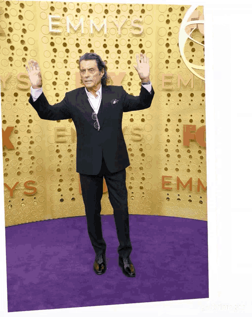 a man in a suit stands on a purple carpet in front of an emmys wall
