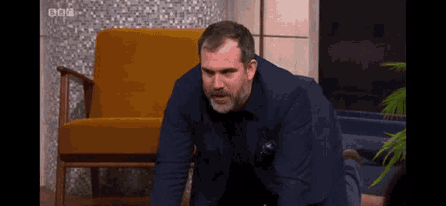 a man in a blue suit is crawling on the floor in front of a yellow chair with bbc written on the bottom