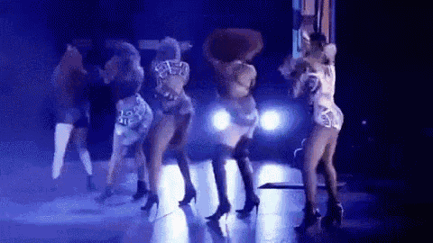 a group of women are dancing on a stage in front of a car in a dark room .