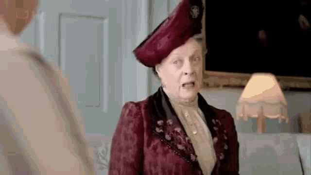 a woman in a red dress and hat is standing next to a lamp and talking to another woman .