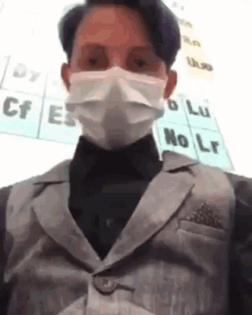 a man wearing a mask and a suit is standing in front of a periodic table of the elements .