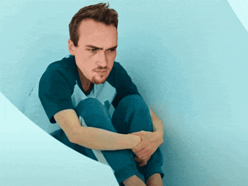a man in a blue shirt and blue pants sits in a corner with his legs crossed