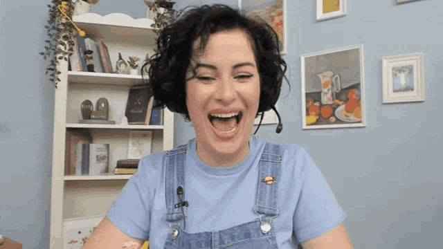 a woman wearing overalls and a blue shirt laughs in front of a bookshelf
