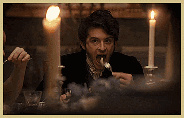 a man sitting at a table with a fork in his mouth and candles in the background
