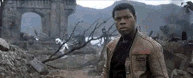 a man in a brown jacket is standing in front of a bridge in a ruined city .