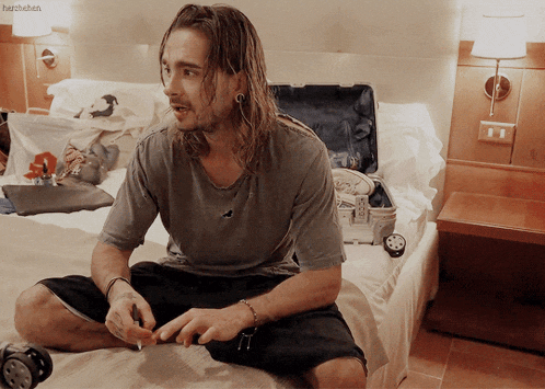 a man with long hair is sitting on a bed next to a suitcase and a lamp with the word herzchen written on it