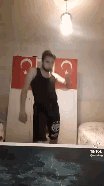 a man in a tank top and shorts is dancing in a room with two flags behind him .