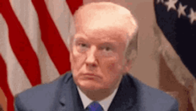 donald trump is sitting in front of an american flag and looking at the camera .