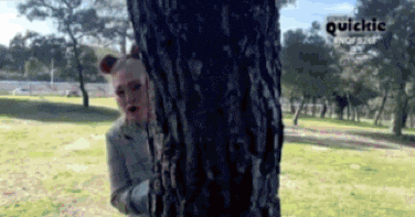 a woman peeking out from behind a tree with quickic written on the bottom right