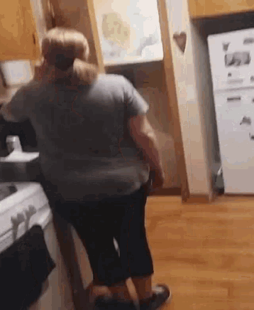 a woman in a gray shirt is standing in a kitchen next to a refrigerator .