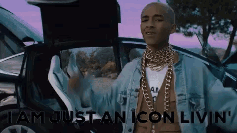 a man wearing a denim jacket and gold chains is standing in front of a car with the door open .