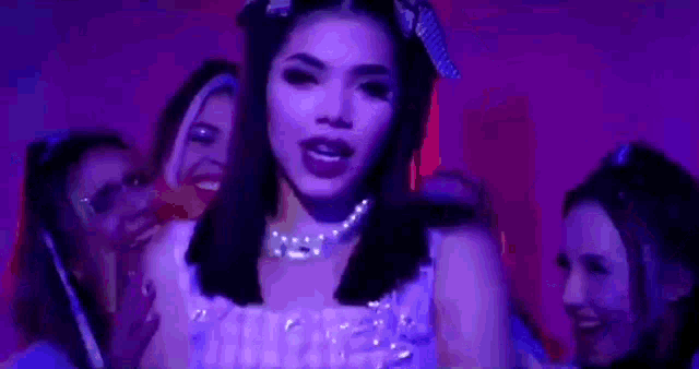 a woman in a purple dress and pearl necklace is surrounded by a group of women in a dark room .