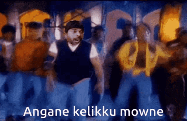 a man in a vest is dancing in front of a crowd with the words angane kelikku mowne written below him