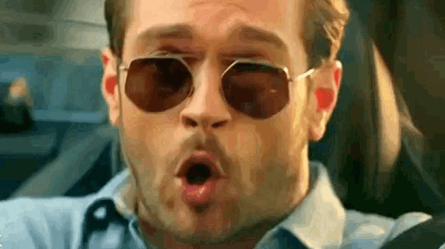 a man wearing sunglasses and a blue shirt is making a funny face while driving a car .