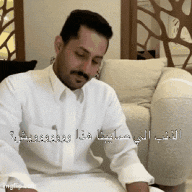 a man in a white shirt is sitting on a couch with arabic writing on his shirt