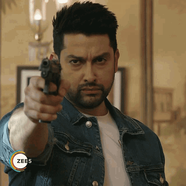 a man in a denim jacket is pointing a gun at the camera with a zee5 logo on the bottom