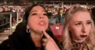 a couple of women are sitting next to each other in a stadium . one of the women is pointing at the camera .
