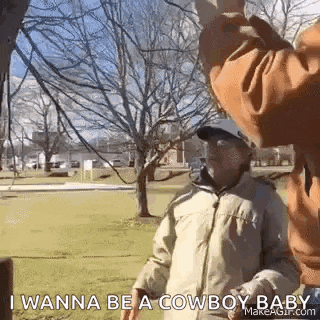 a man is standing in a park talking to another man while wearing a cowboy hat .