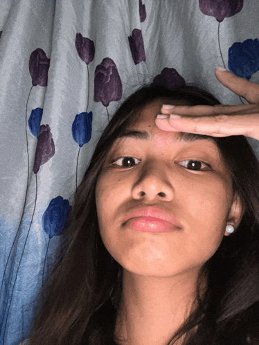 a girl with a ring on her finger touches her forehead in front of a curtain with purple and blue flowers