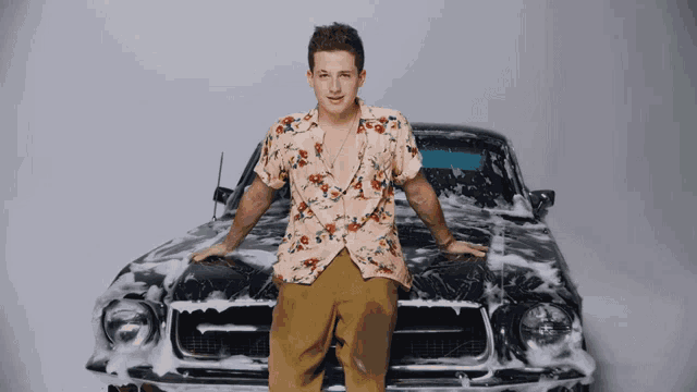 a man in a floral shirt leans on the hood of a car covered in soap
