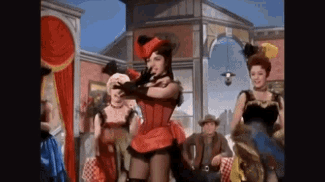 a woman in a red corset and gloves is dancing in front of a crowd of people in a western town .