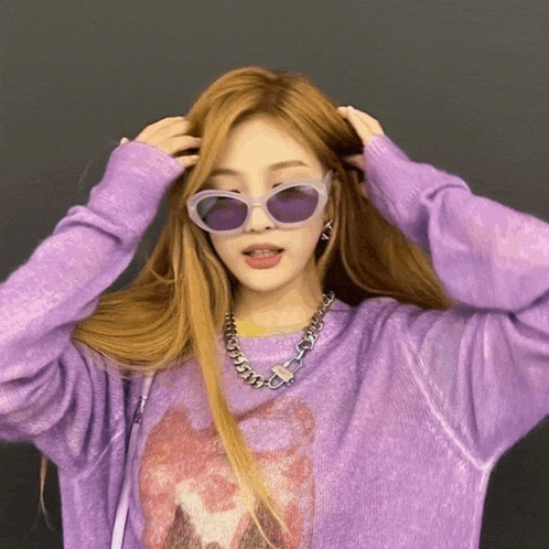 a woman wearing sunglasses and a purple sweater holds her hair
