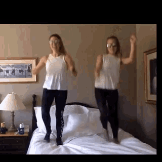 two women wearing sunglasses are jumping on a bed in a room