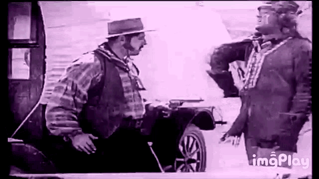 two men are standing next to each other in front of a car and talking .