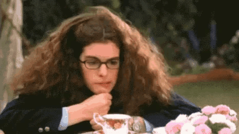 a woman with long hair and glasses is sitting at a table drinking from a teacup .