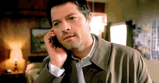 a man in a trench coat and tie is talking on a cell phone in a living room .